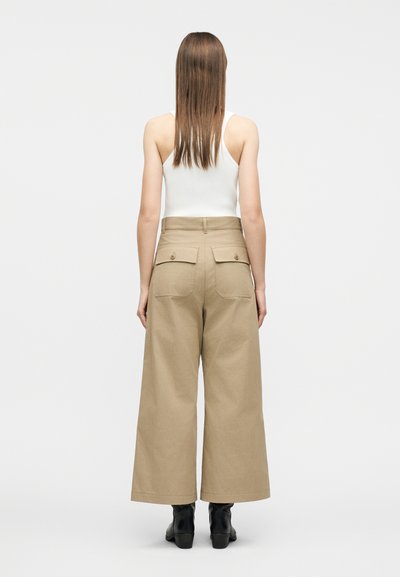 Γυναίκα με ίσια καστανά μαλλιά που φοράει λευκό τοπ χωρίς μανίκια, παντελόνι με ευρείες γραμμές σε μπεζ χρώμα με πίσω τσέπες και μαύρες μποτάκια μέχρι τον αστράγαλο, στέκεται με την πλάτη προς τα εμπρός.