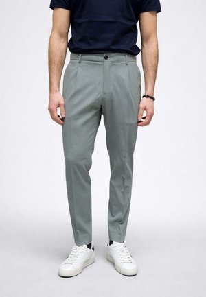 Uomo che indossa pantaloni sartoriali grigio chiaro, sneakers bianche, braccialetto nero e camicia a maniche corte blu navy, in piedi su uno sfondo chiaro e uniforme.