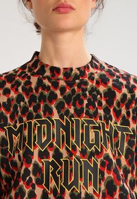 Femme portant un haut imprimé léopard avec des taches rouges, noires et vertes, arborant un texte audacieux entouré de jaune « MIDNIGHT RUN » sur la poitrine.
