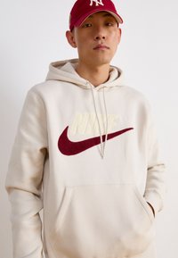 Beige huvtröja med en framficka, prydd med en stor krämfärgad och vinröd Nike-logotyp. Mjuk tyg med dragsko i huvan. Röd keps på huvudet.