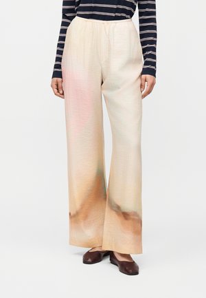 Femme portant un pantalon beige à jambes larges avec un dégradé doux marron et rose, associé à des chaussures plates marron foncé et un haut à manches longues rayé.