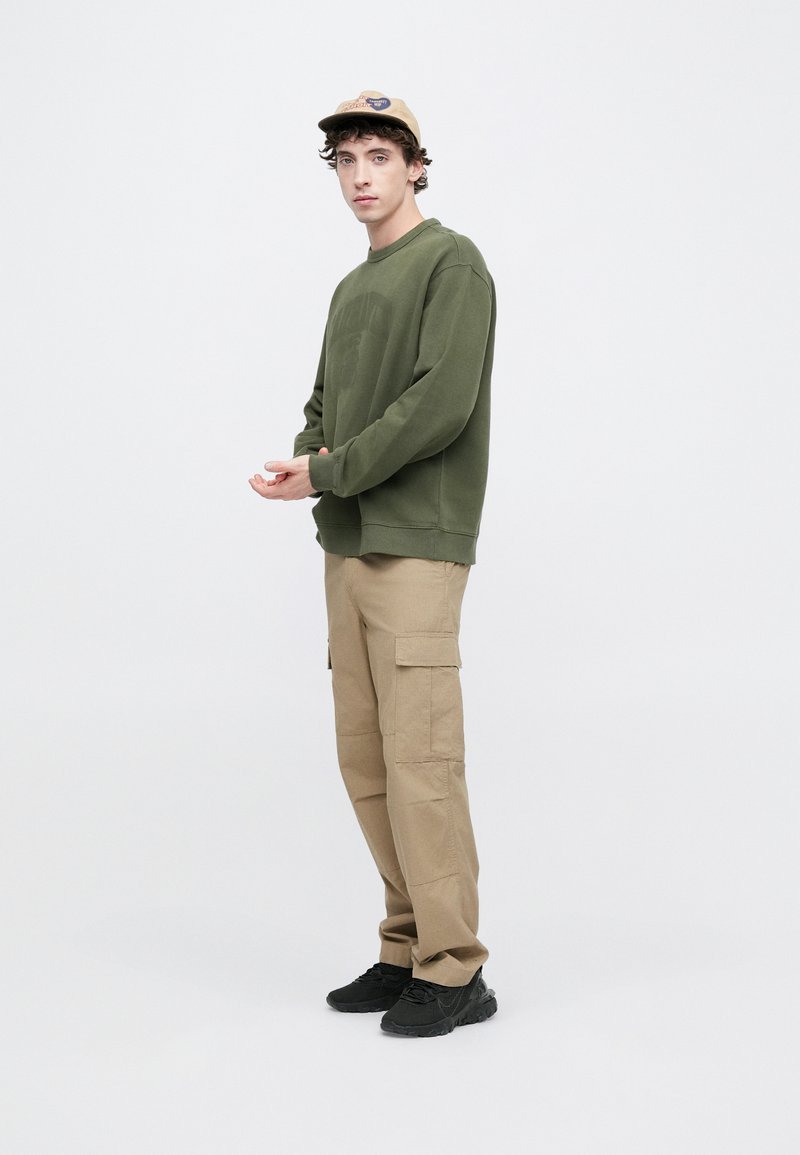 Green cotton sweatshirt, beige cargo pants with side pockets, black sneakers, tan cap with logo, standing against a plain background.