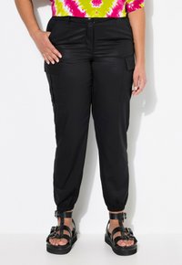 Schwarze Cargo-Hosen mit elastischen Beinabschlüssen, ausgestattet mit Seitentaschen und einem Reißverschluss. Kombiniert mit schwarzen Sandalen mit Riemen auf einem weißen Hintergrund.