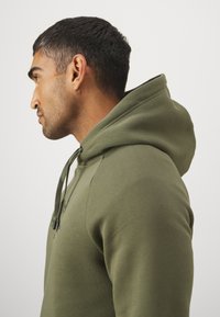 Olivengrøn hættetrøje med en glat tekstur, tætsiddende design og snore. Har side- og skuldersømme, som viser en moderne silhuet.