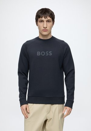Jeune homme portant un sweat-shirt noir à manches longues avec l'inscription "BOSS" sur le devant et un pantalon beige, debout devant un fond blanc uni.