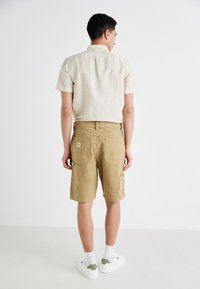 Bermudashorts og stripete kortermet skjorte i lyse beige nyanser, laget av bomull, med en avslappet passform og to baklommer. Hvite sneakers.