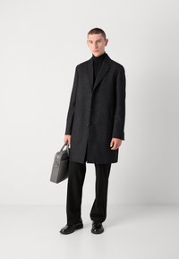 Cappotto lungo in misto lana nera con una silhouette strutturata, design a un petto e una texture sottile. Abbinato a pantaloni neri e calzature.