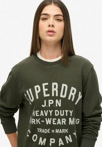 Sudadera verde oliva con cuello redondo y mangas largas. Presenta texto impreso en blanco, incluyendo "SUPERDRY" en letras gruesas. Textura suave.