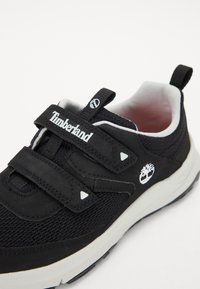 Crna sportska cipela s mrežastim gornjim dijelom, dva podesiva Velcro remena, bijelim detaljima i teksturiranom gumenenom potplatom. Timberland logo sa strane.