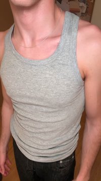 Canotta grigia a costine senza maniche con scollo rotondo, abbinata a jeans neri. Una sottile catena d'argento è visibile attorno all'area del collo.