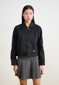 Veste noire courte avec col, fermeture éclair dorée et écusson. Portée par-dessus une jupe plissée grise. Texture lisse et design structuré.