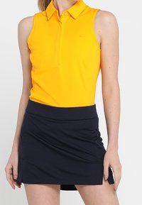 Gul ärmlös pikétröja med krage kombinerad med en marinblå skort. Piken har en diskret logga och en mjuk tygtextur.