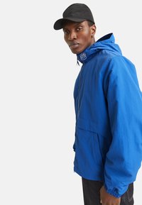 Leichte blaue Jacke mit Kapuze, ausgestattet mit einer Fronttasche, elastischen Bündchen und einem Kordelzug am Kragen. Strukturierter Stoff.