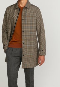 Mężczyzna ubrany w brązowy, kraciasty płaszcz na guziki, noszony na rudym pomarańczowym swetrze i ciemnoszarych plisowanych spodniach, stoi z jedną ręką w kieszeni.