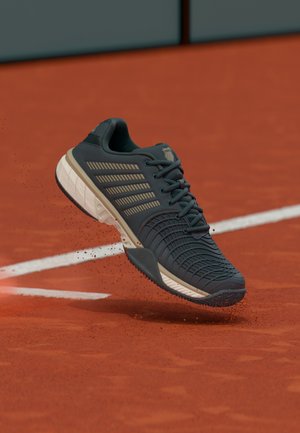 Sportska cipela tamnosive boje s bež detaljima podiže prašinu od gline na crvenom teniskom terenu blizu bijele granice.