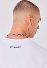 Hombre con corte de cabello corto tipo buzz y una cadena de plata, vistiendo una camiseta blanca con "PXP" impreso en la parte posterior, de perfil.