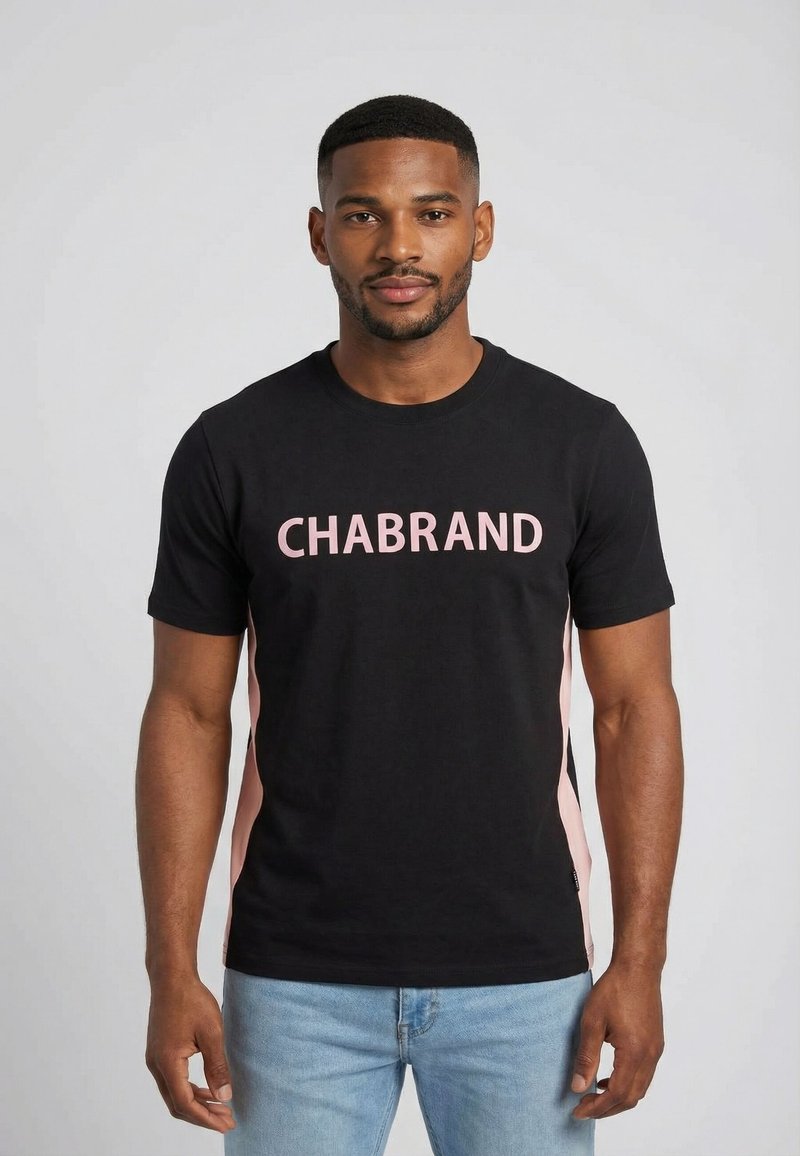 Hombre con camiseta negra con paneles laterales rosas y texto "CHABRAND" en rosa, combinado con vaqueros azul claro, de pie frente a un fondo liso.