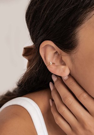 Gold hoop earring with a small decorative accent. Close-up view of the earlobe, showing skin tone and textured hair against a neutral background.