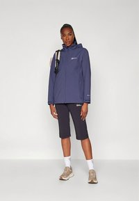 Giacca impermeabile blu navy con cappuccio, chiusura frontale con zip e logo. Abbinate a pantaloncini neri fino al ginocchio e sneaker beige, in piedi su uno sfondo semplice.