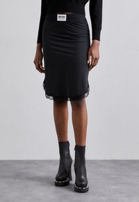 Kvinna i en svart Moschino Jeans kjol med spetsdetaljer, svart långärmad topp och svarta klackade ankelstövlar med kraftig sula.