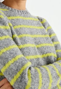 Harmaa neulepaita, jossa on kirkkaan keltaisia vaakaraitoja, pyöreä kaula-aukko ja teksturoidusta, pehmeästä kankaasta.