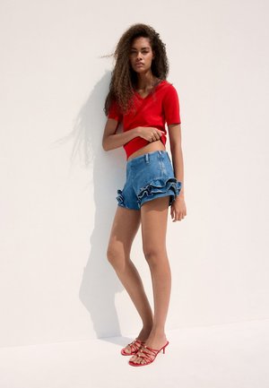 Jeune femme aux longs cheveux bouclés, portant un haut rouge à manches courtes, un short en jean à volants et des sandales rouges à talons avec brides, debout contre un mur blanc.