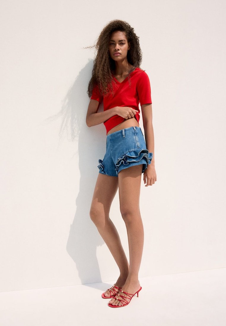 Jeune femme aux longs cheveux bouclés, portant un haut rouge à manches courtes, un short en jean à volants et des sandales rouges à talons avec brides, debout contre un mur blanc.