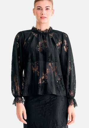 Femme portant un chemisier en dentelle noire à manches longues avec un col à volants et une jupe en dentelle assortie sur fond blanc.
