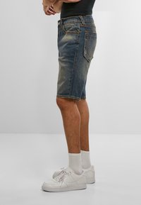 Osoba v bledých denimových šortkách, bielych ponožkách po členky a bielych športových topánkach, stojaca pred jednoduchým svetlosivým pozadím.