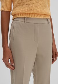 Beige maßgeschneiderte Hosen mit Fronttaschen und einer glatten Textur, kombiniert mit einem gelben Strickpullover mit kurzen Ärmeln.