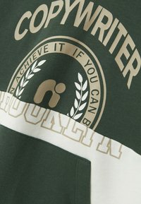 Sweatshirt vert arborant un grand motif "COPYWRITER", des accents dorés et une bande blanche contrastante avec le texte "BROOKLYN". Matière douce, design décontracté.