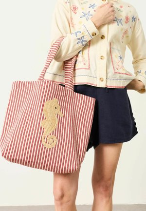 Woman wearing embroidered cream jacket and navy shorts, holding red-and-white striped tote bag with woven seahorse design.