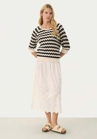 Un pull à motif zigzag noir et blanc associé à une jupe blanche légère avec un ourlet festonné et un design à œillets. Porté avec des sandales beiges.