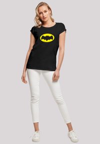 Black cotton T-shirt featuring a large yellow Batman logo on the front. Paired with white skinny pants and white sneakers.