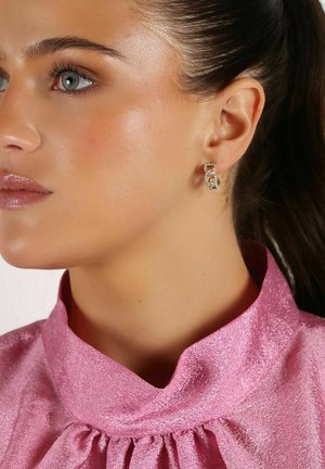 Guldtonade örhängen med en länkad design och klara stenar som dekoration, bärs av en person i en rosa, strukturerad topp med hög krage.
