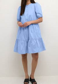 Ljusblå bomullsklänning med korta puffärmar, rund halsringning, knappstängning fram, lager-på-lager nederdel och avslappnad passform. Svarta klacksandaler.