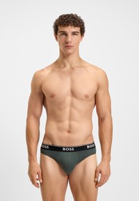 Boxeri scurți de culoare verde închis, cu un tiv negru pe care este inscripționat numele brandului "BOSS" cu litere albe. Textură netedă și design ajustat.