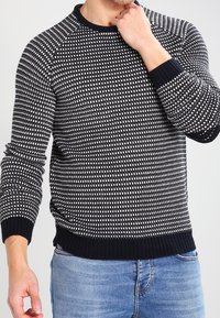 Pull en tricot présentant un motif à pois noir et blanc, col rond, poignets et ourlet côtelés, assorti à un jean bleu clair.