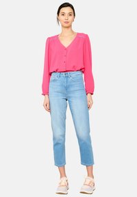 Blusa rosa de manga larga con botones en el frente y hombros fruncidos, combinada con jeans recortados de color azul claro y zapatillas multicolor.