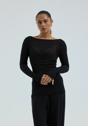 Femme aux cheveux tirés en arrière, portant un haut noir ajusté à manches longues et un pantalon noir ample, debout les mains jointes devant un fond clair uni.