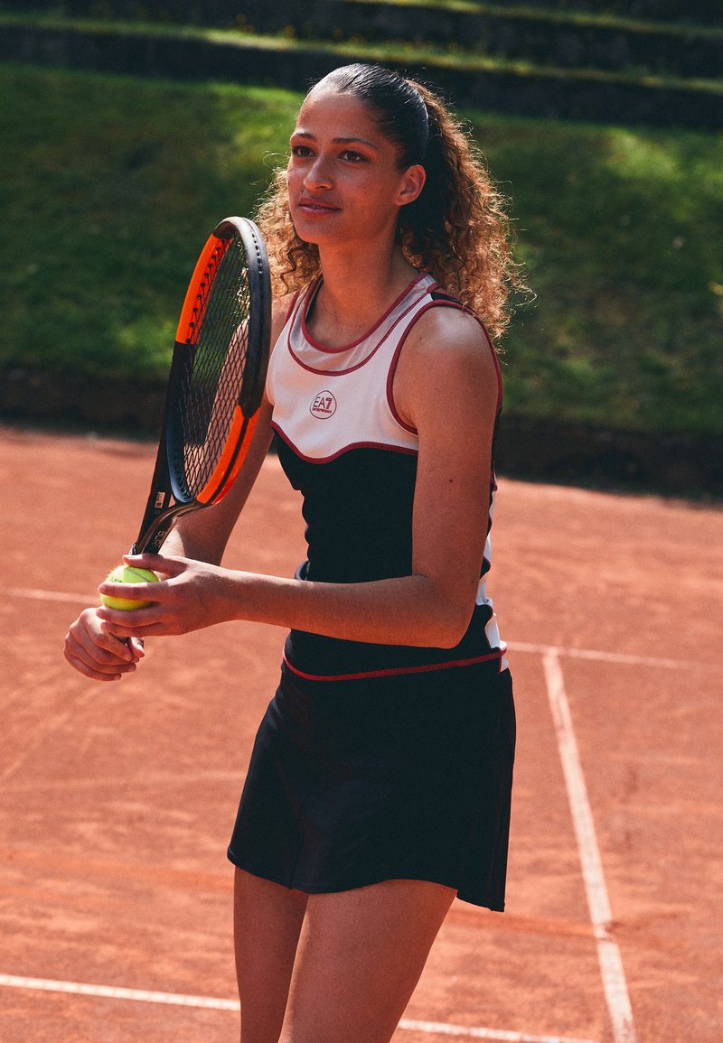 Atleta feminina vestindo um traje de ténis em preto e branco, segurando uma bola de ténis amarela e uma raquete, posicionada num court de terra batida.