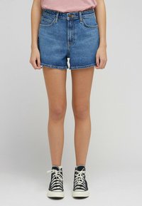 Pantalones cortos de mezclilla en azul claro, de cintura alta con un dobladillo deshilachado. Combinados con zapatillas negras de caña alta que tienen cordones blancos y suelas de goma.