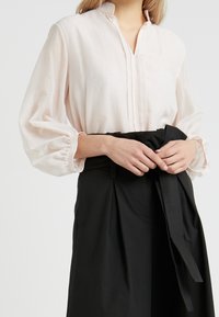 Blouse rose clair à manches longues avec un détail plissé à l'avant, associée à une jupe noire taille haute avec une taille nouée et une texture de tissu douce.