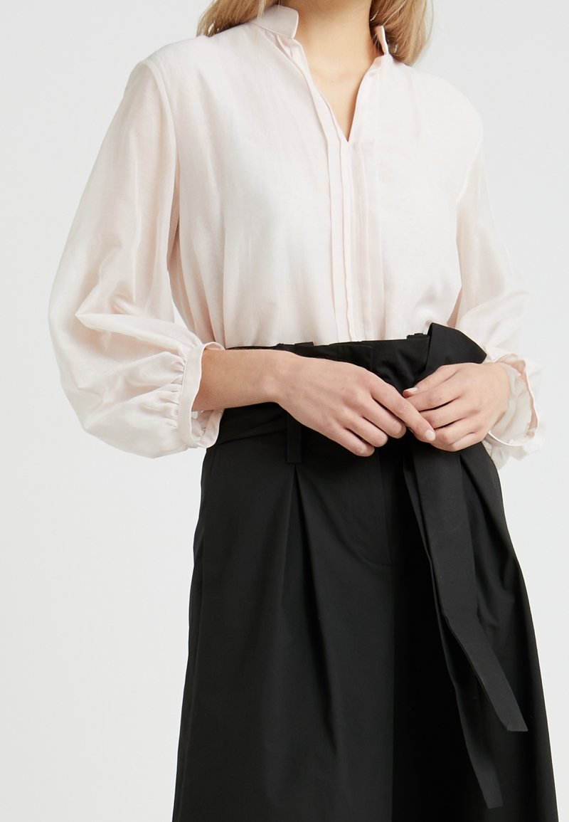 Blouse rose clair à manches longues avec un détail plissé à l'avant, associée à une jupe noire taille haute avec une taille nouée et une texture de tissu douce.