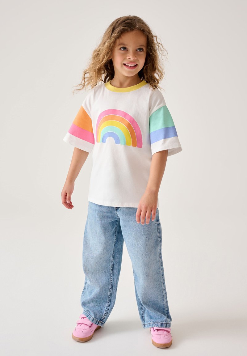 Valge lühikese varrukaga T-särk, millel on vikerkaare graafika roosa, oranž, kollane, roheline ja sinine, kombineeritud helesiniste lõdva lõikega teksade ja roosade kingadega.