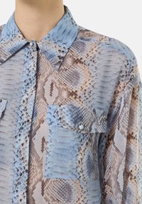 Camicia leggera e traslucida con un motivo a pelle di serpente blu e beige. Include un colletto con bottoni e una tasca frontale.