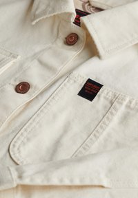 Chaqueta de mezclilla blanca con botones de cobre, que presenta un bolsillo frontal y una etiqueta de marca roja y negra. La textura es suave con una apariencia resistente.