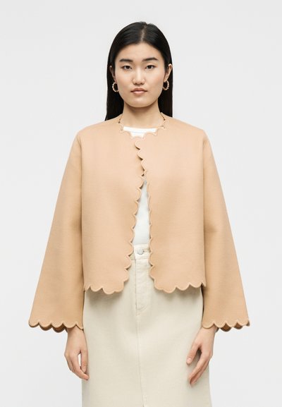 Γυναίκα με ίσια σκουρόχρωμα μαλλιά φοράει μπεζ σακάκι με κυματιστό τελείωμα πάνω από λευκό τοπ και φούστα κρεμ ψηλόμεση, στέκεται μπροστά σε μονόχρωμο φόντο.