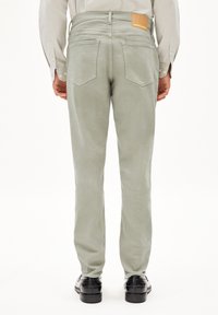 Pantalons en velours côtelé vert clair, ajustés, avec une texture lisse, cinq poches et une étiquette en cuir marron à la taille, portés avec des chaussures noires.