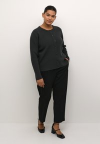 Dunkelgrauer gestrickter Cardigan mit langen Ärmeln und vorderem Knopfverschluss, kombiniert mit schwarzen Hosen und schwarzen Blockabsatzschuhen.